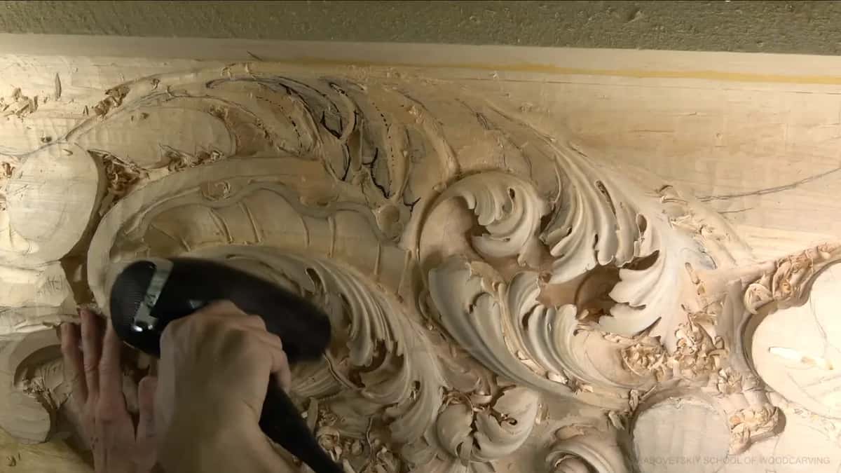 A black hair dryer, held close in both hands, pushes chips away from the carved leaves.