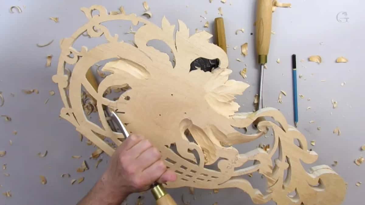 A hand pushes a gouge into the back of the panel while more gouges and a blue pencil lie on the right