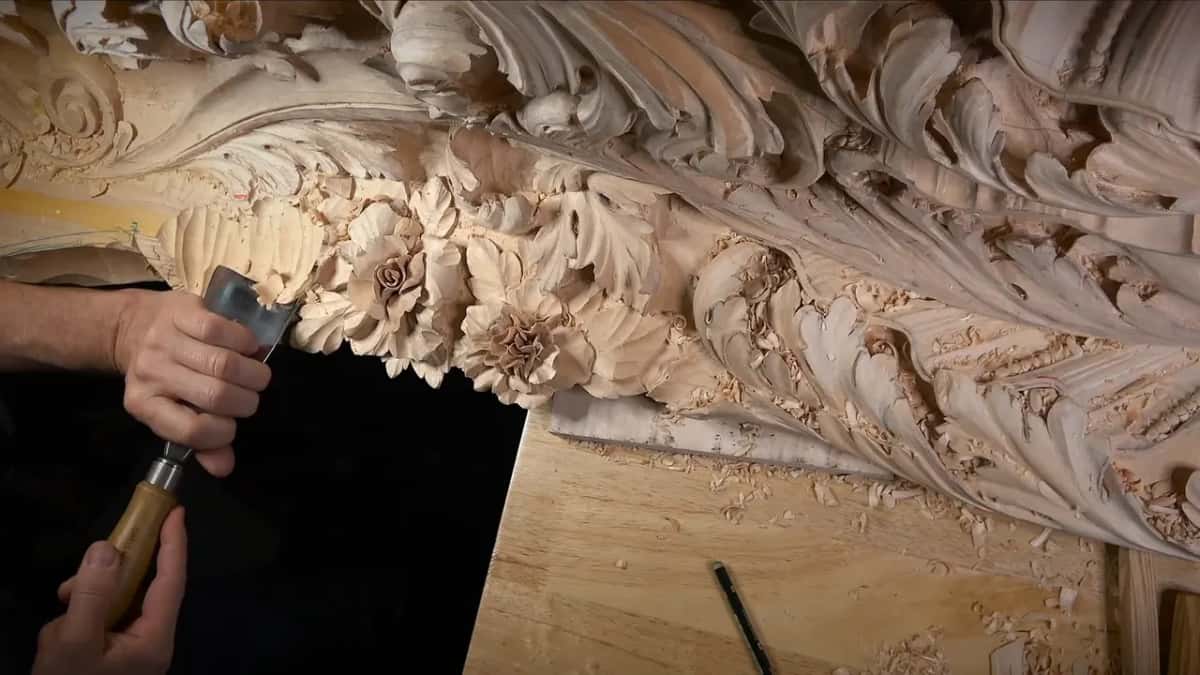 Both hands drive a large gouge along the panel's edge beside the finished flowers