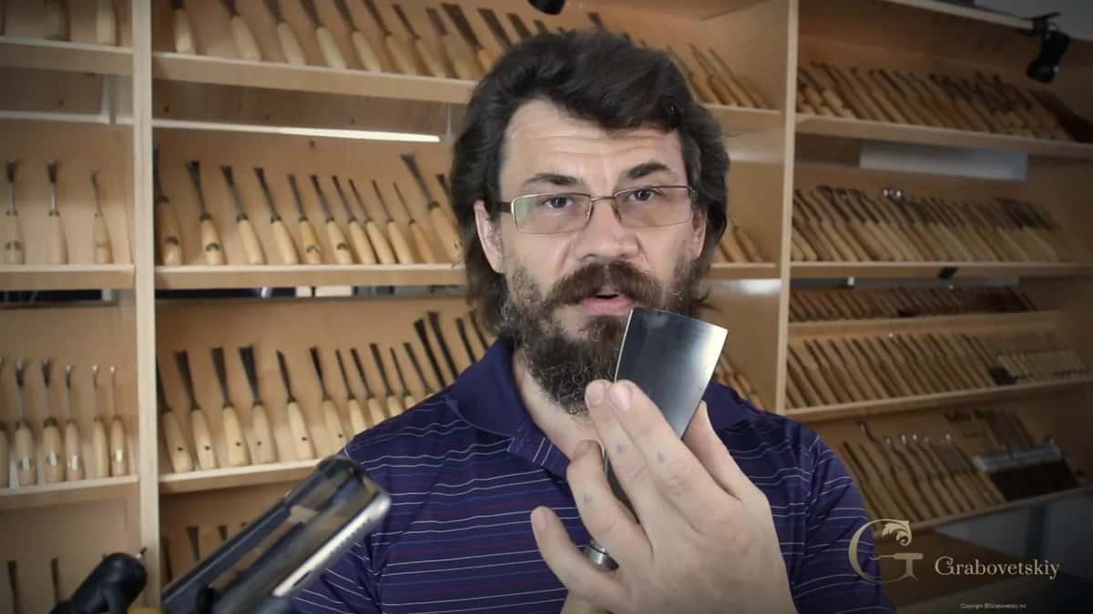 Alexander holds the wide #7 blade up beside his face, the shop wall behind him packed with rows of carving gouges.
