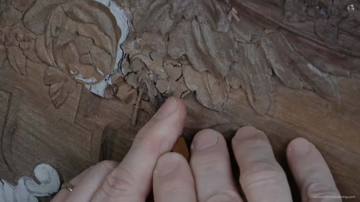 Fingers flat on the carving, a pale handled gouge setting the direction the petal travels in.
