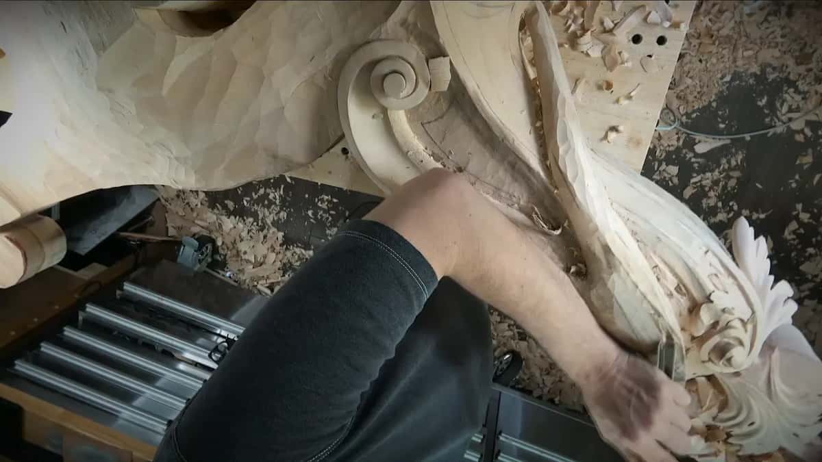 One hand steering a long gouge down the band from above, curls of shavings piled by the scroll