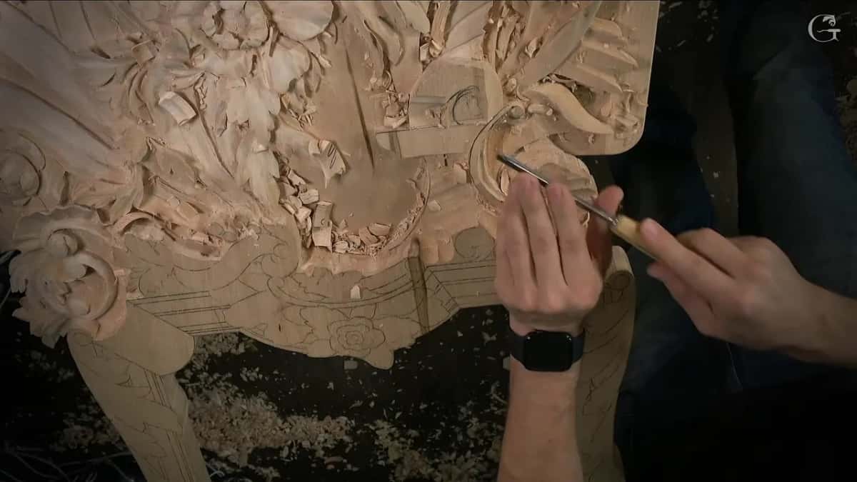 Seen from above, two hands guide a slim gouge around the volute while chips gather below the molding