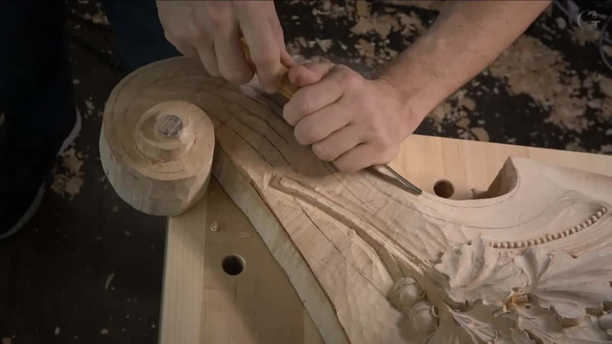 Two hands push one gouge along the top edge of the scroll, pencil grid lines crossing the wood below.