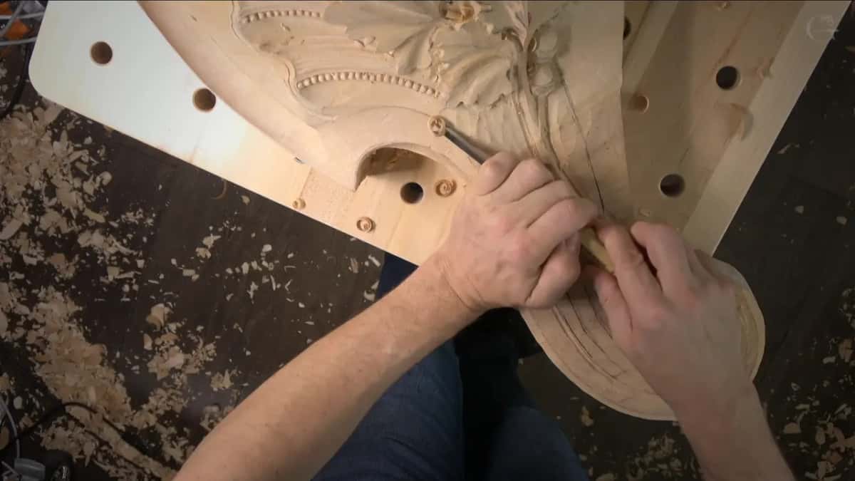 Both hands guide a narrow veiner through the curve, bench holes and dark shavings below.
