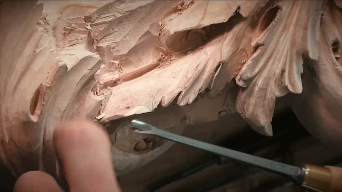 Brass-ferruled gouge sliding in beneath the carved leaves, a fingertip at the left edge of the picture