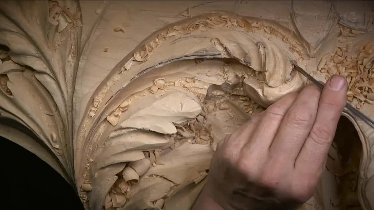 A thin tool held like a pencil at the edge of a carved leaf, shavings all around