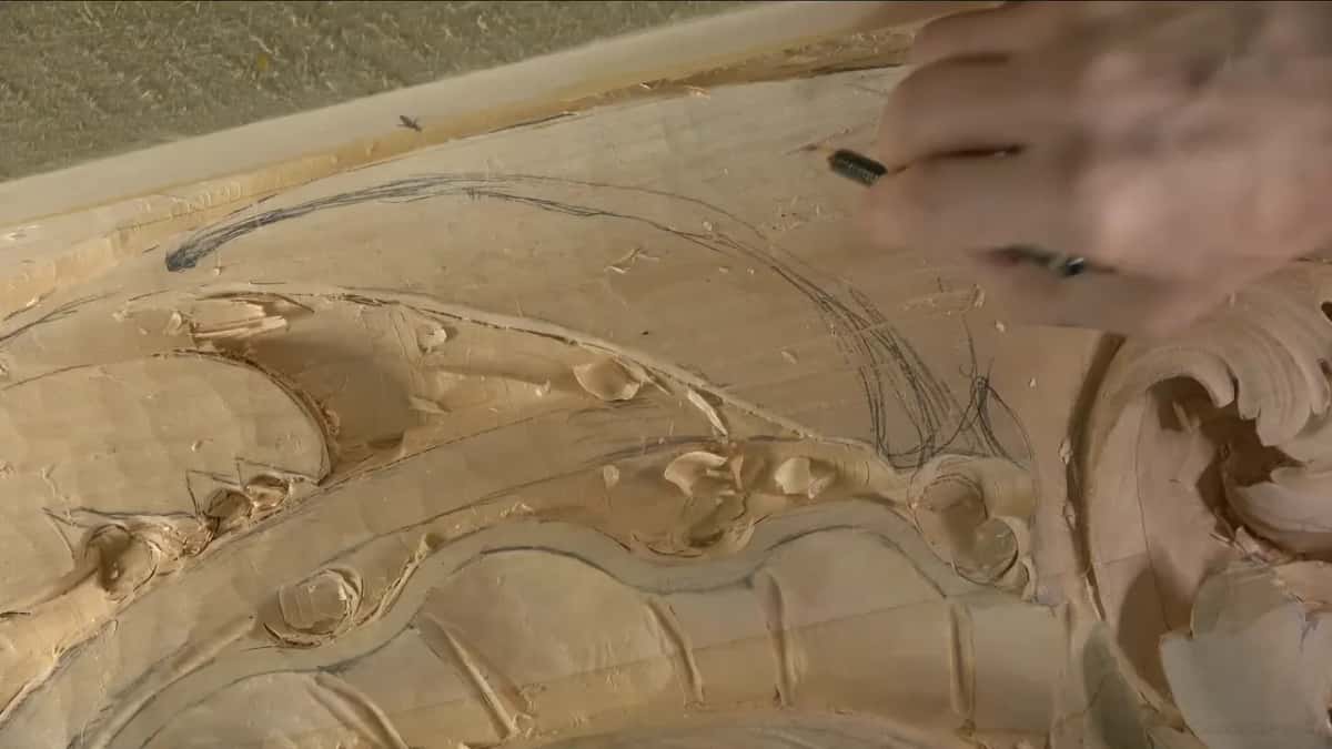 Pencil in hand, he sketches loose curves on the uncarved wood above the carved leaves.