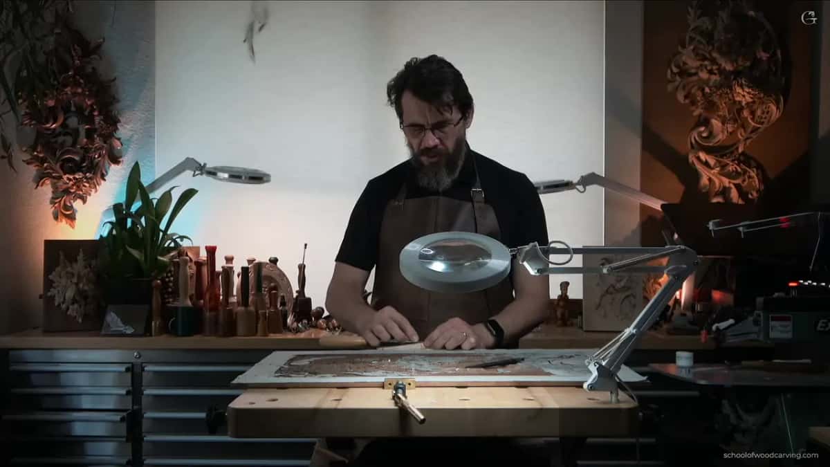 Across the shop, him under the magnifier lamp with the panel flat in front of him and jars of tools behind.