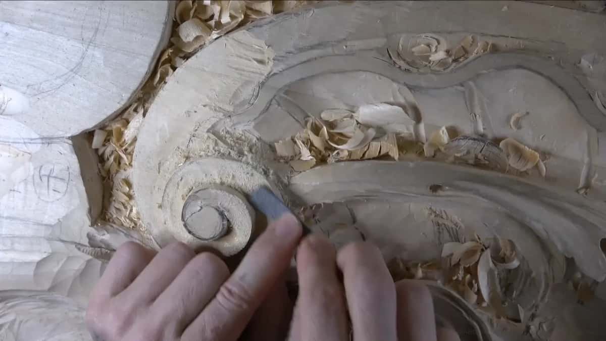Two hands meet at the volute's rim, one pressing a small gray rasp flat, the other's fingers resting beside it.