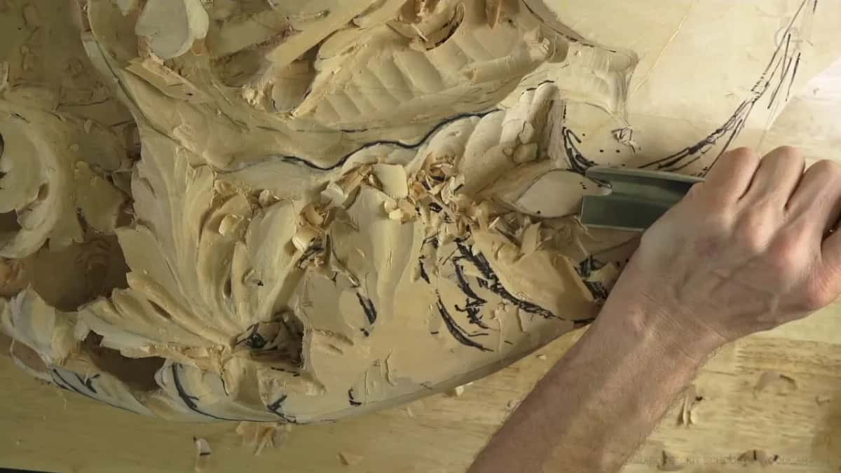 One hand drives a wide gouge into the right edge, beside a spill of fresh chips.