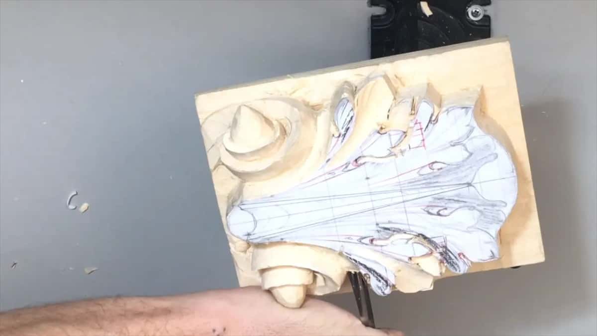 Board turned on its side, volute at the left, one hand low on the bottom edge holding a short tool