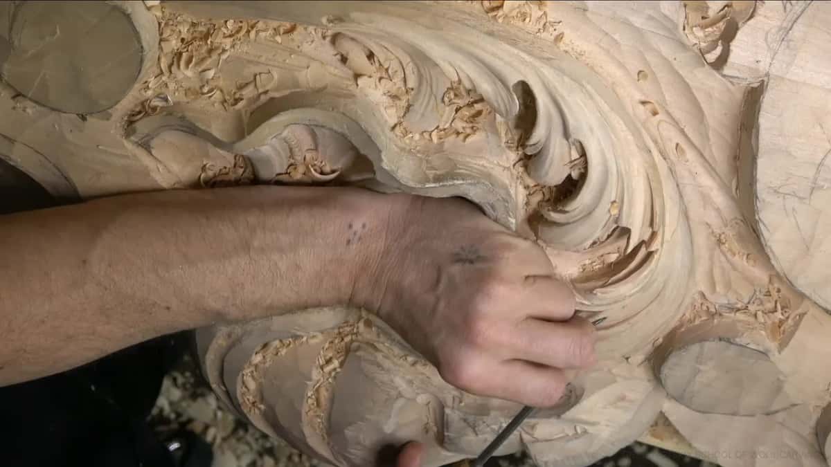 Knuckles over a curling lobe, the tool tip hidden in its hollow, shavings scattered across the shell.