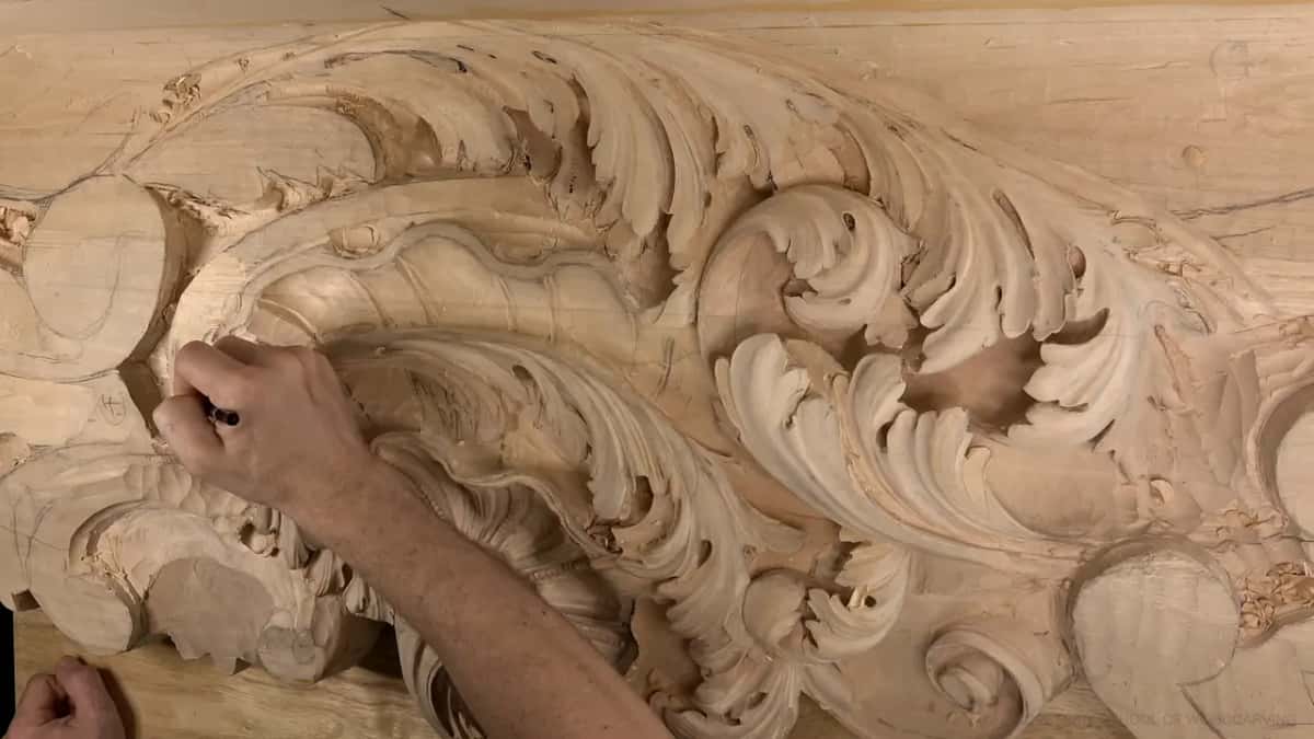 Fingers grip a thin black tool by the volute, acanthus curls filling the right.
