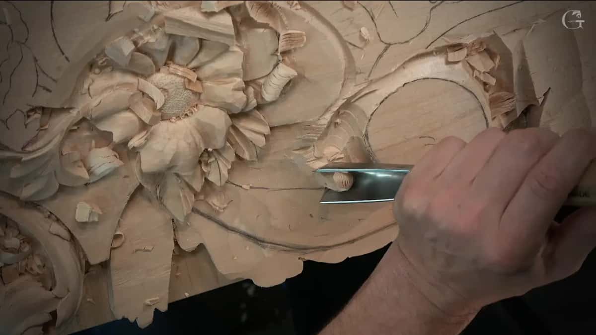 A wide gouge in one hand lowers the wood between the flower and the round scroll