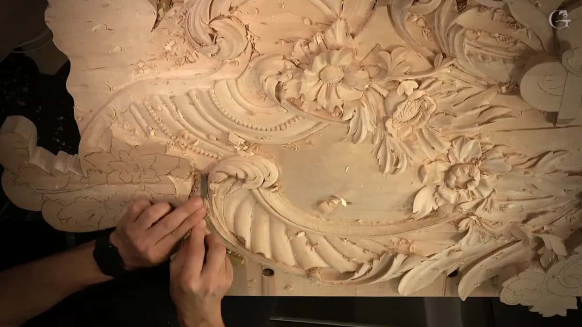 Two hands on a tool, far left, carved flowers across the top of the panel