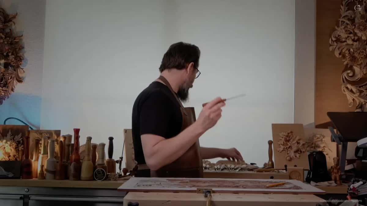 Side view across the workshop, tool in hand, carvings hanging behind.