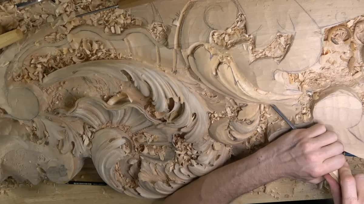 One hand steers a slim gouge at the right edge of the finished head, the rough leaf beyond it.