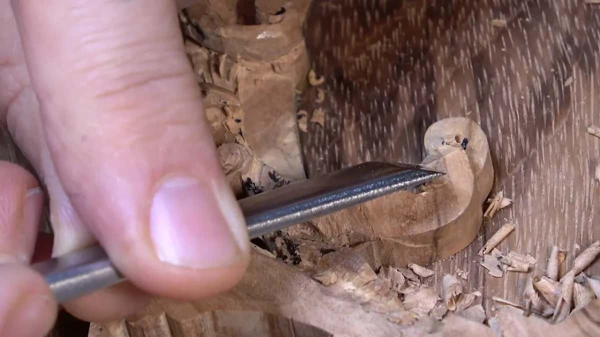 A gouge lies low across the first scroll's end while a thumb rides on its shaft, shavings piled to the right