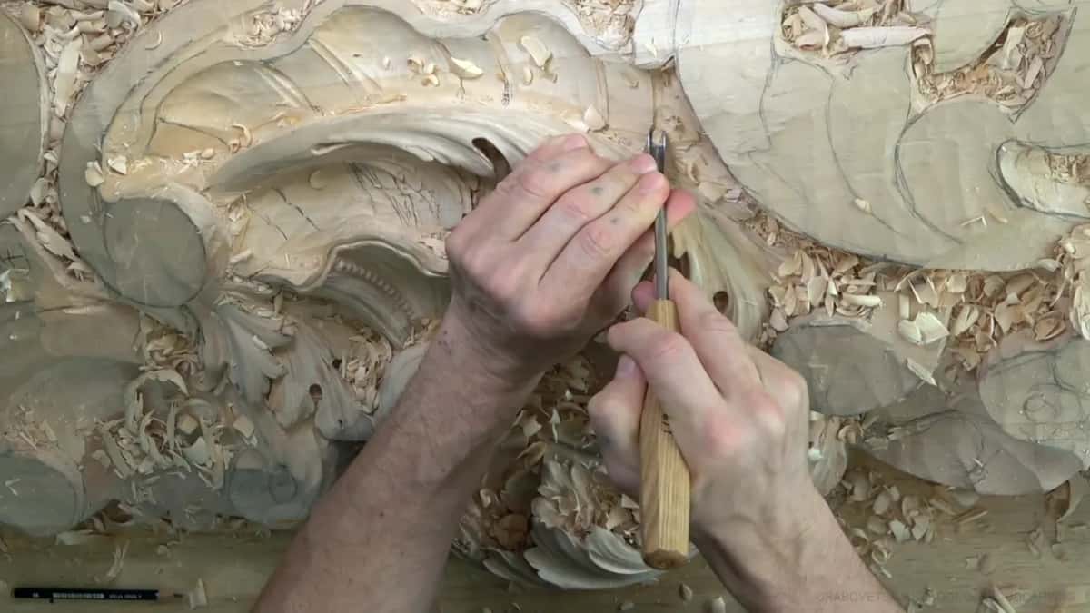 Both hands guide a narrow gouge under the big scroll, beside the round shell.
