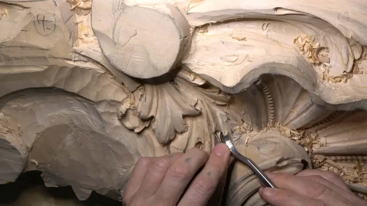 Two hands at work, one steadying while the other guides a bent spoon gouge into a hollow near the scroll.