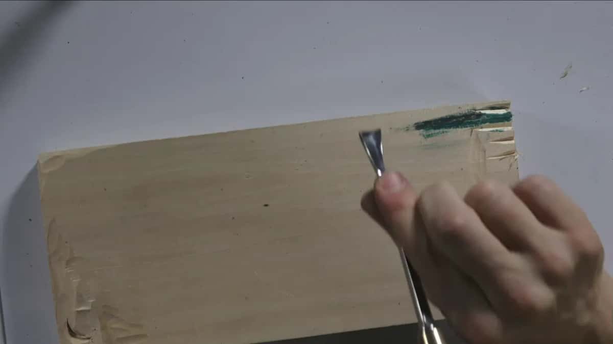 Pale block with green compound smeared at its top edge, a hand drawing a gouge along it.