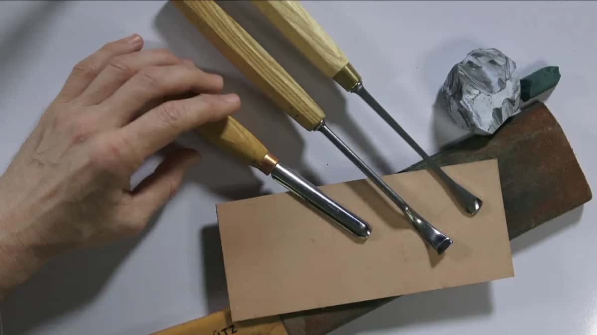 Three carving tools lie across a piece of tan leather, with a foil lump and a green one behind them.
