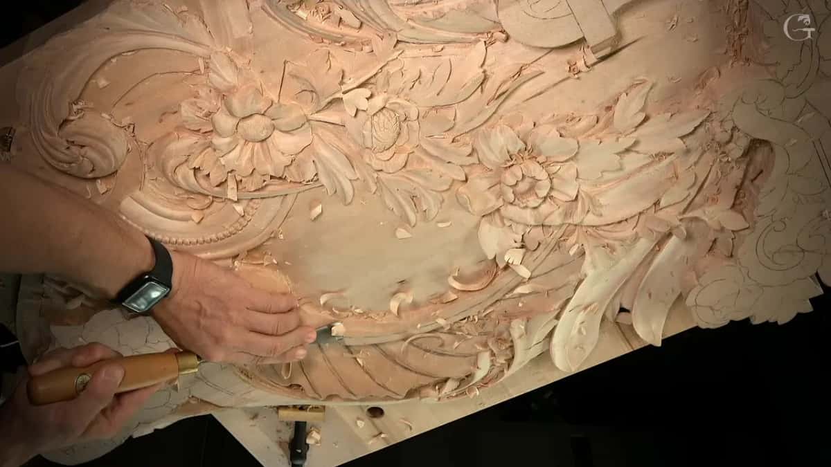 From above, one hand steers a gouge along the lower curve while three carved flowers sit above