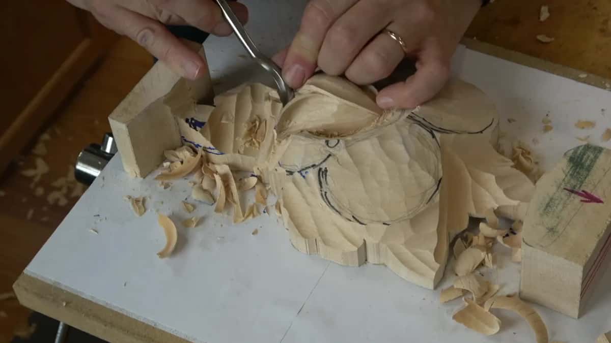 Two hands on a spoon gouge at the left side of the panel, one shaving still attached