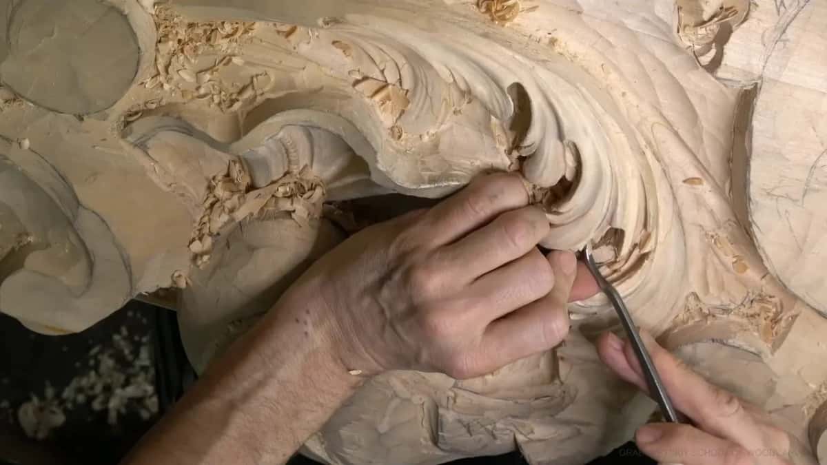 Both hands work a small tool under the thin edge of an overhanging curl from its left side.