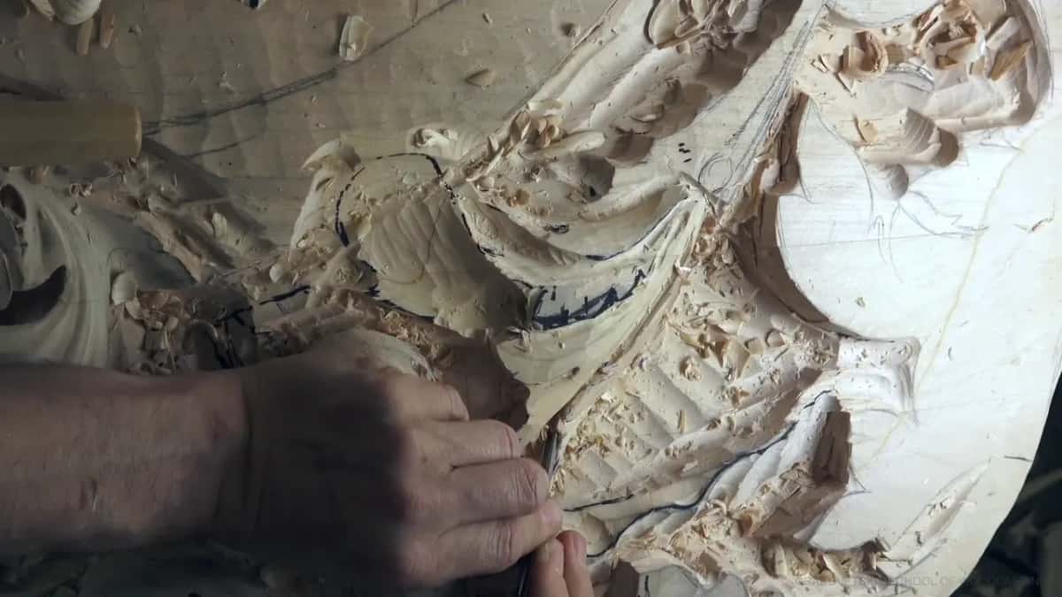 A thin tool works low under a curl, among dozens of blue marker lines on pale wood.