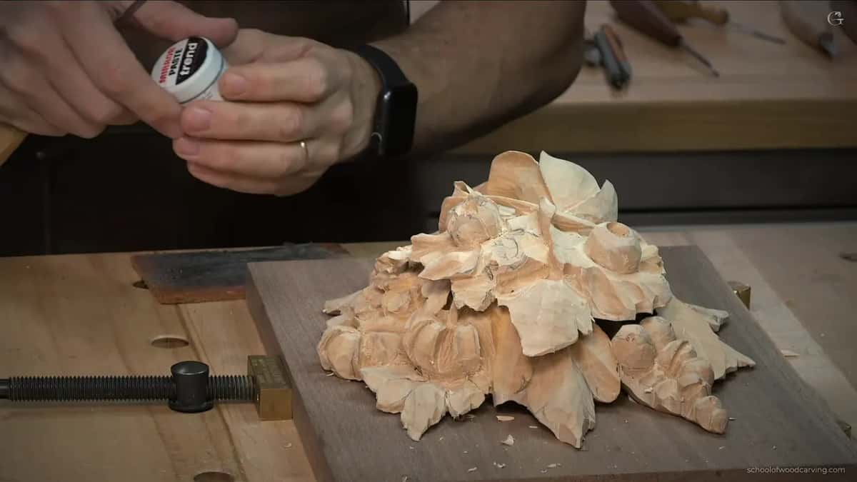 A small tin of paste held up over the carving on its walnut board