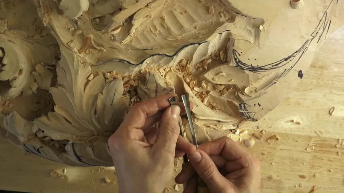 Two gouge blades pinched upright between the fingers of both hands, side by side, just above the carved leaves.