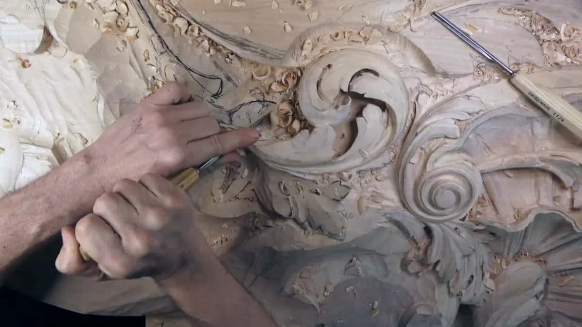 Both hands work below the scroll, a spare gouge lying on the board above.
