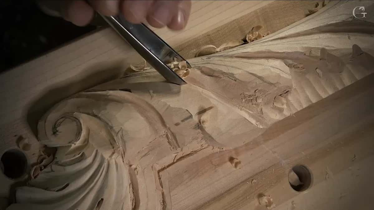Wide flat chisel reaching in under the edge of a carved leaf, shavings at its tip