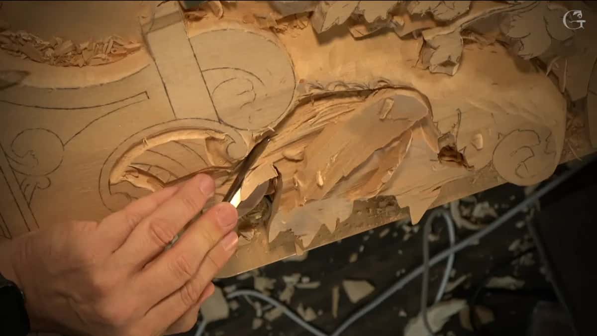Fingers hold a narrow gouge down in a curved groove beside the lowered part.