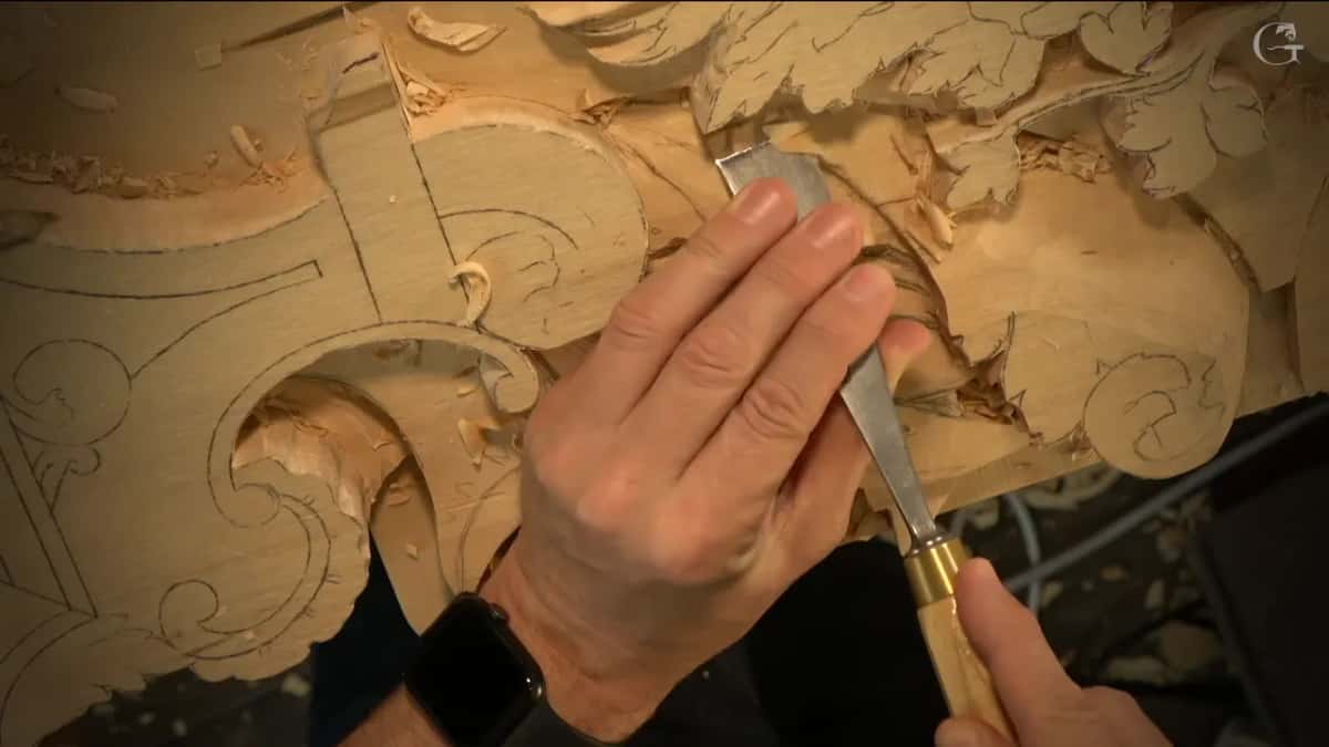 Fingers holding a flat chisel against a carved scroll.