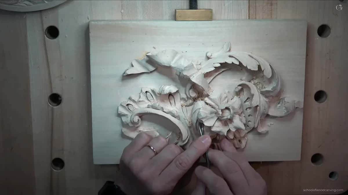 Overhead, both hands guiding a thin blade into the flower of a carved rocaille panel that lies flat on the bench.