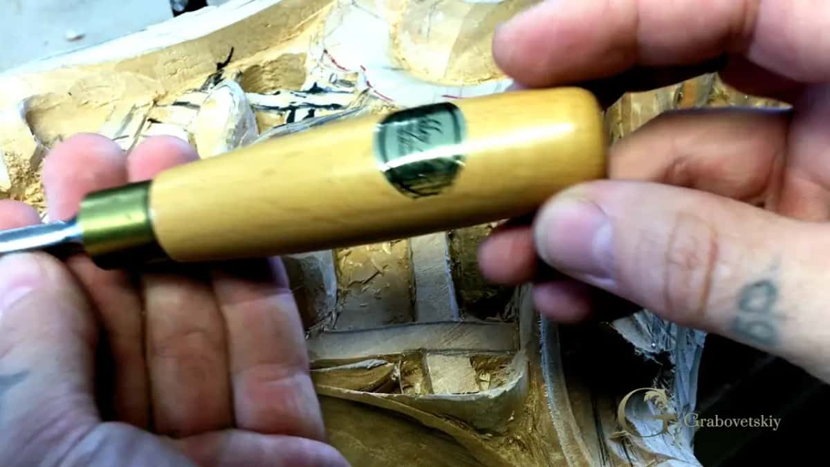 Close on a pale handle with a green oval maker stamp, held over a carving that has already been gilded.
