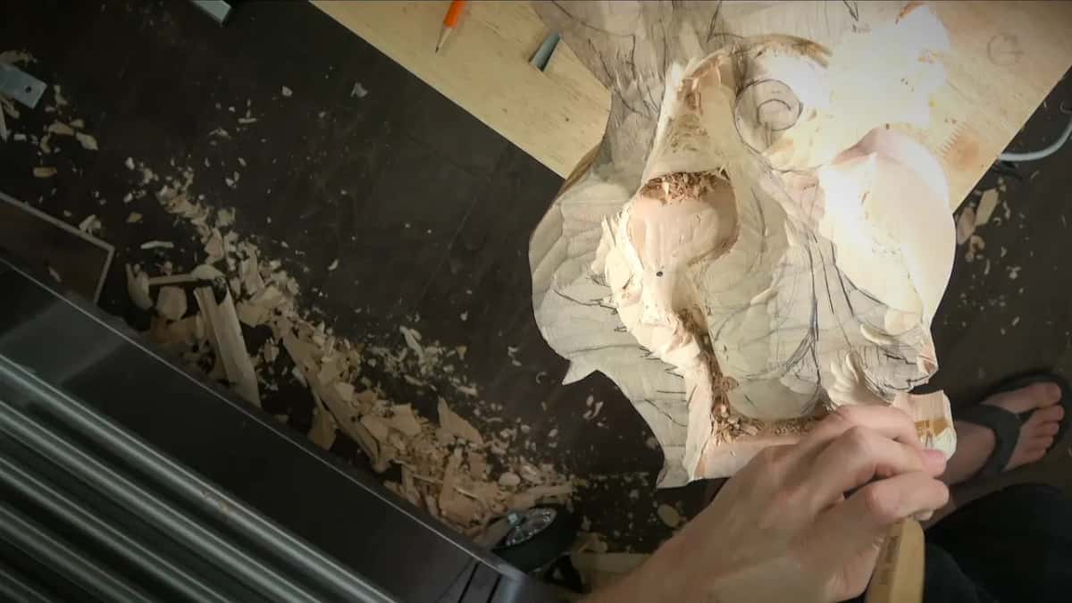 Hands grip a gouge at the base of the carving