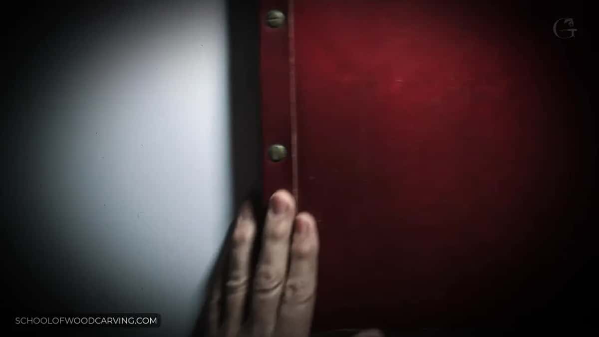 A hand opens the red cover of the antique catalog, two brass studs at its edge