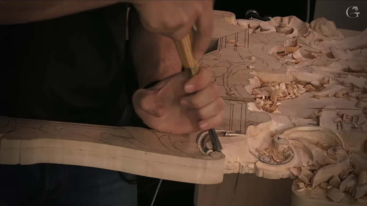 A narrower gouge gripped in two hands low on that same edge, pencil lines all over the board