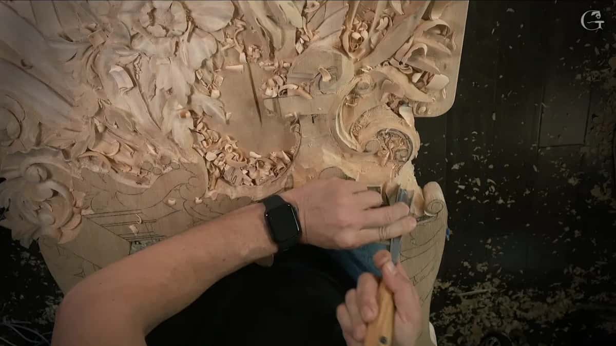 Two hands work a gouge at the right edge of the panel
