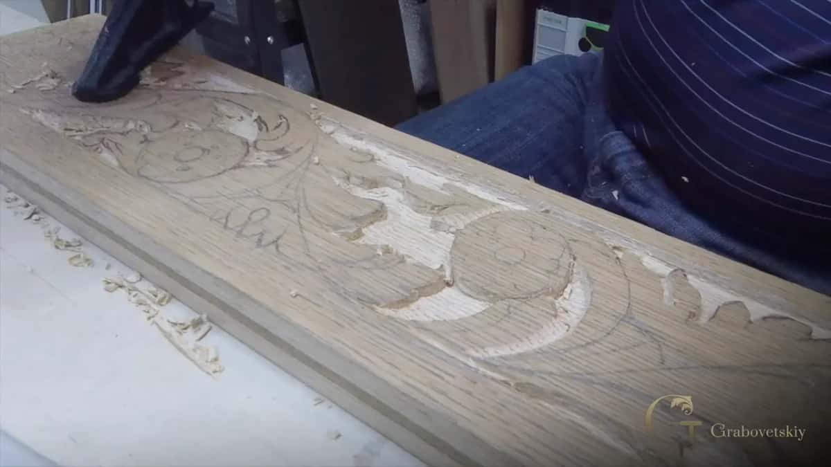 An apron board with its scrolls half carved, held under a blue clamp at the far end