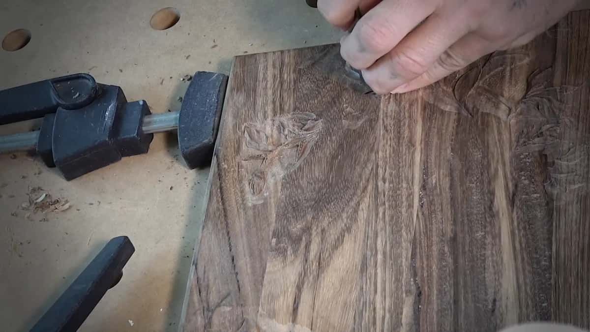 Fingers press a punch into dark wood right beside the carving, a black clamp at the edge.