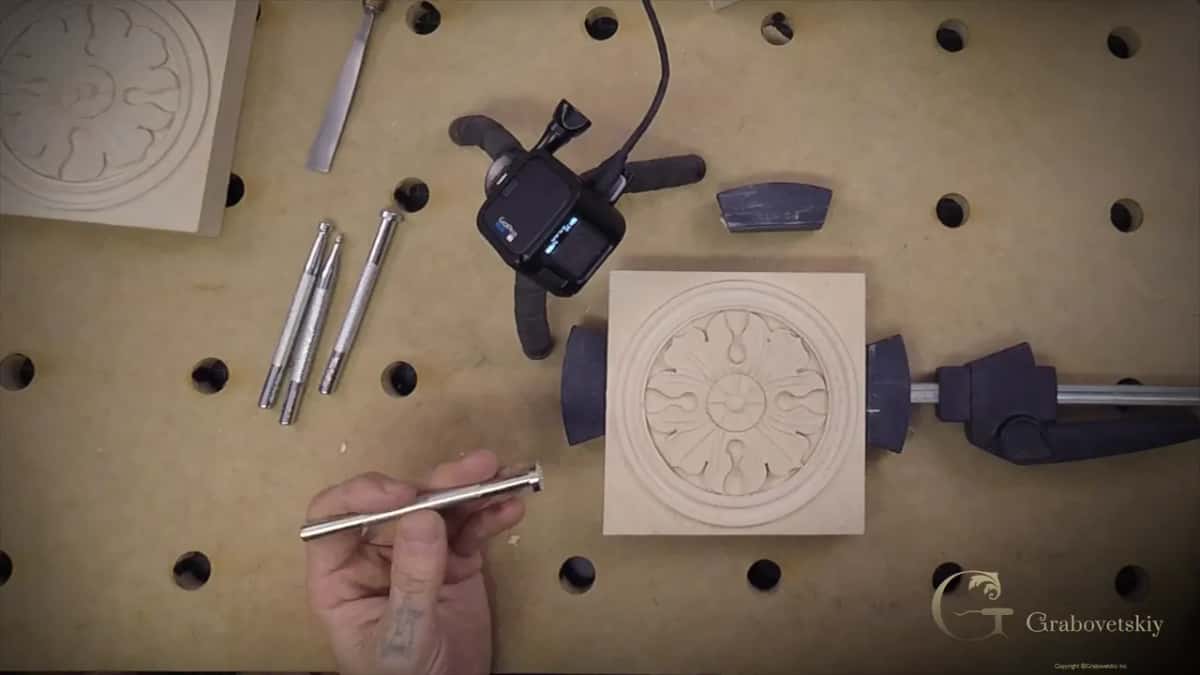 A slim metal punch held up beside the clamped rosette, with three more resting near a mallet.