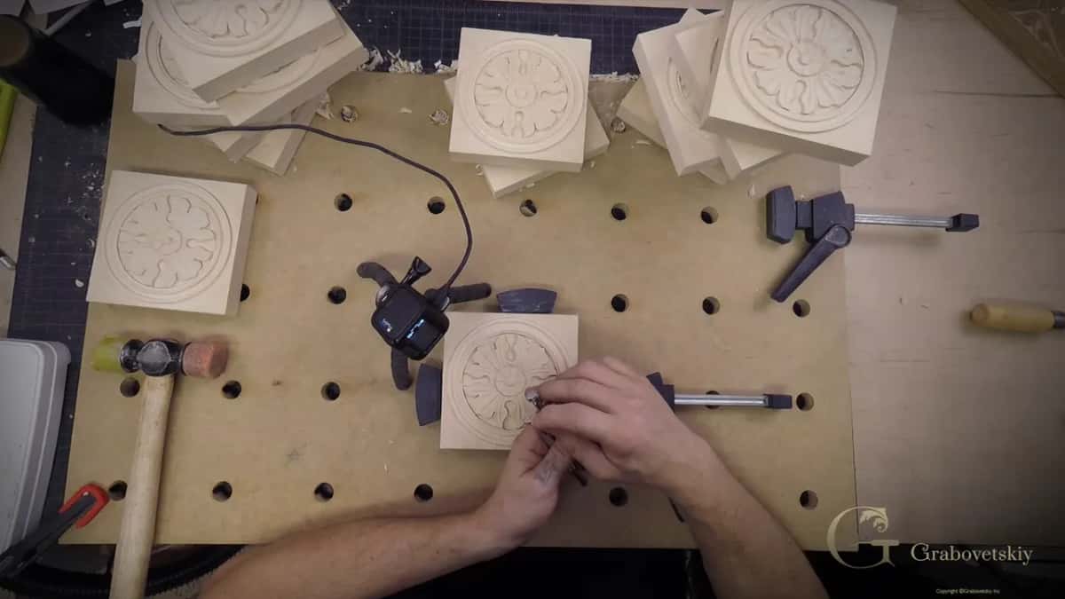 Hands at a clamped rosette block on a drilled bench, with finished rosettes stacked behind.
