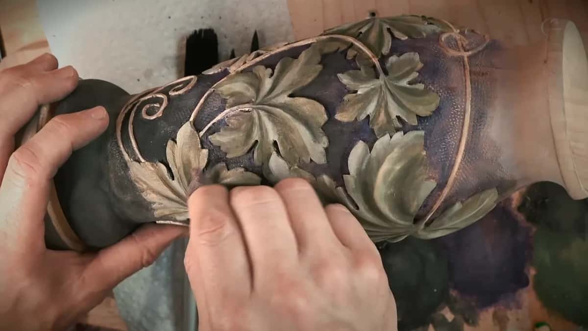 Both hands turn the colored vase, its leaves green and gold over a purple ground with pale vines