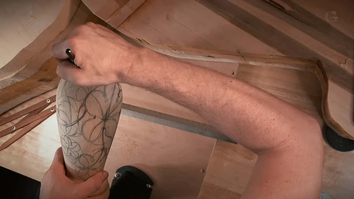A hand sketches grape leaves in pencil across the bare turned vase before any carving