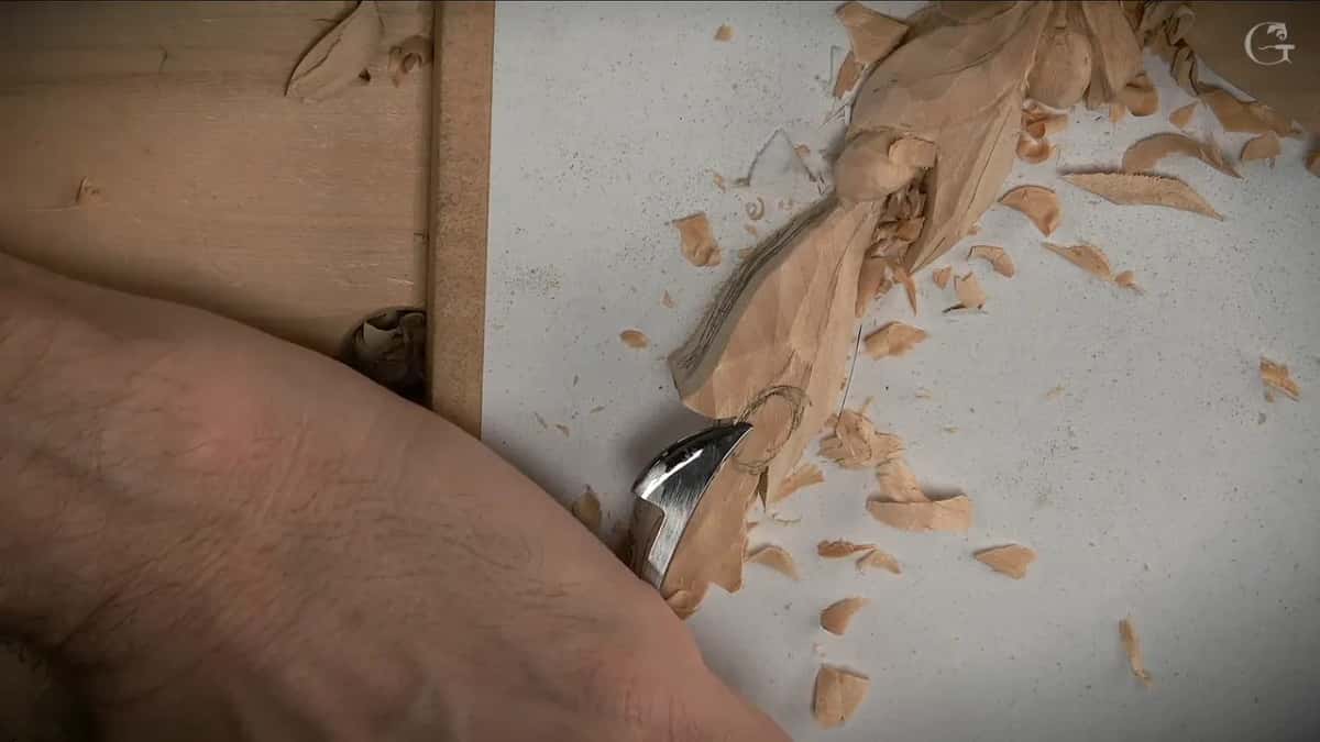 Bent gouge resting against a leaf being shaped, loose shavings scattered on the white board
